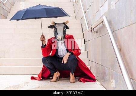 Geschäftsfrau mit Bullenmaske und Regenschirm, während sie sich in der Nähe der Treppe hockend Stockfoto