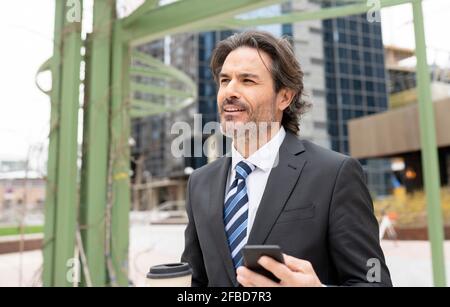 Lächelnder männlicher Profi, der wegschaut, während er die Kaffeetasse hält und Mobiltelefon Stockfoto