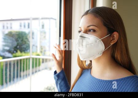 Nahaufnahme eines Portraits einer Frau, die im Innenbereich im Gesicht steht Maske KN95 FFP2 schaut durch das Fenster Stockfoto