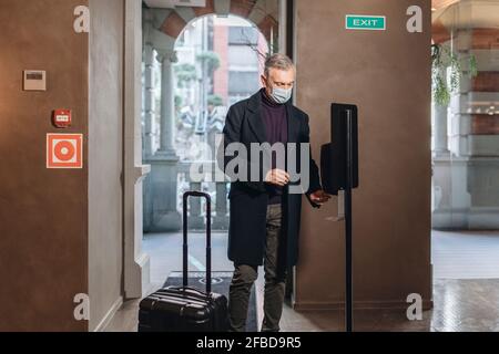 Mann mit Gesichtsschutzmaske, der im Stehen ein Händedesinfektionsmittel anwendet Mit dem Gepäck im Hotel Stockfoto