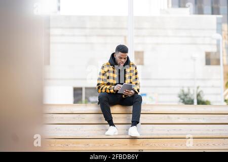 Hübscher junger Mann mit digitalem Tablet, während er auf Holz sitzt Bank Stockfoto