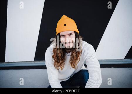 Lächelnder Mann mit langen Haaren sitzt auf dem Fußweg Stockfoto