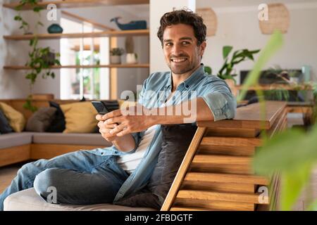 Lächelnder Mann schaut weg, während er das Smartphone zu Hause hält Stockfoto