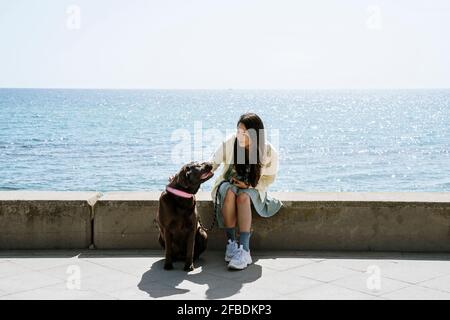 Frau, die mit dem Labrador-Hund auf der Stützmauer vor der Tür sitzt Des Meeres Stockfoto