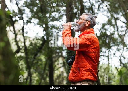 Älterer Mann mit Kamera, der Wasser trinkt, während er im Wald steht Stockfoto