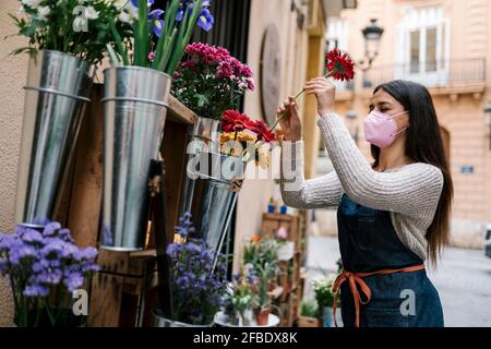 Junge weibliche Floristin mit schützender Gesichtsmaske arrangiert Gerbera Daisy Blume im Laden Stockfoto