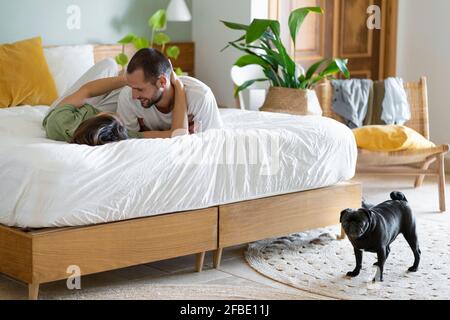 Glückliches junges Paar umarmt auf dem Bett, während Pug Hund stehend Zu Hause auf dem Boden Stockfoto