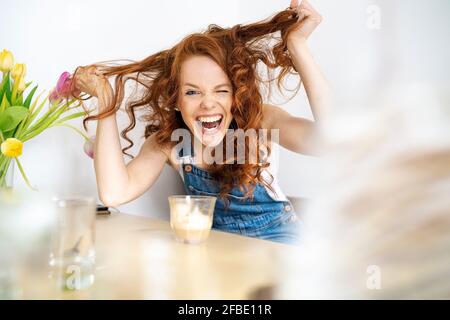 Portrait von Fröhliche Frau Haar ziehen während der Sitzung mit Getränk am Tisch im Zimmer Stockfoto