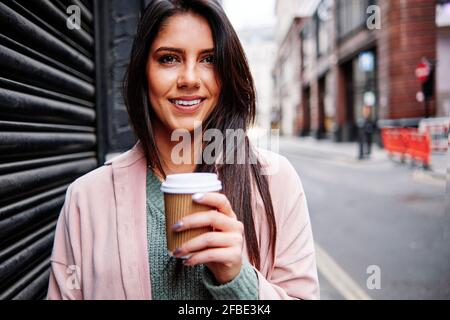 Lächelnde junge Frau mit Einweg-Kaffeetasse, die beim Verschluss steht In der Stadt Stockfoto