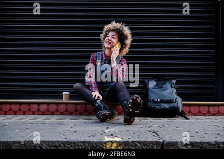 Lächelnde, lockige Frau, die im Sitzen auf dem Mobiltelefon spricht Auf Fußweg Stockfoto