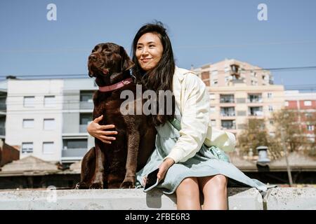 Lächelnde Frau, die mit Chocolate Labrador auf der Stützmauer sitzt Vorderseite des Gebäudes Stockfoto