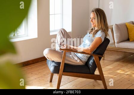 Lächelnde blonde Frau schaut weg, während sie mit einem digitalen Tablet sitzt Auf dem Sessel zu Hause Stockfoto