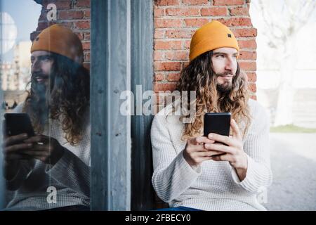 Bärtiger Mann mit Mobiltelefon schaut weg, während er sitzt Vorderseite der alten Mauer Stockfoto