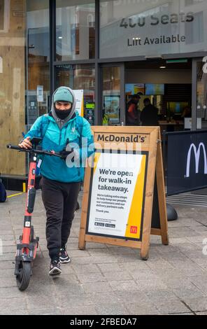 Deliveroo Bike-Lieferkurier mit Gesichtsmaske, der McDonald's mit einer Lebensmittelbestellung verlässt. Auf einem gemieteten Elektroroller geliefert werden. Stockfoto