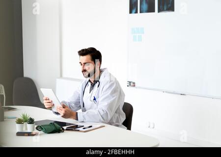 Hübscher männlicher Arzt mit Tablet, der im Krankenhaus am Schreibtisch sitzt Stockfoto