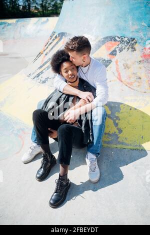 Junger Mann küsst sich auf die Stirn der Freundin, während er am Skateboard sitzt parken Sie an sonnigen Tagen Stockfoto