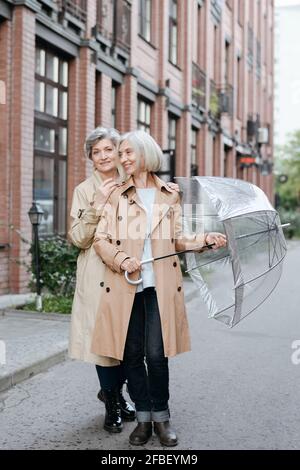 Lächelnde reife Freundinnen mit Regenschirm auf der Straße Stockfoto