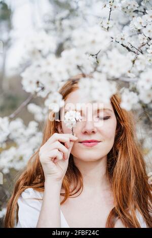 Schöne Frau hält weiße Blume vor dem Auge, während Inmitten eines Mandelbaums stehen Stockfoto