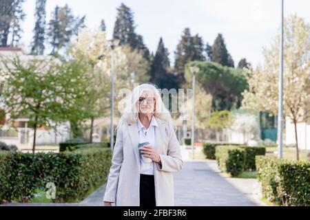 Lächelnde Frau, die mit einer wiederverwendbaren Kaffeetasse auf dem Weg steht Stockfoto