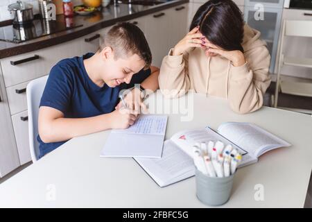 Frustrierte Mutter, die dem Sohn beim Erledigen der Hausaufgaben hilft, während sie darauf sitzt Esstisch zu Hause Stockfoto