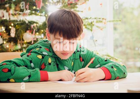 Netter Junge, der am Tisch mit Weihnachtsbaum auf Papier schreibt Im Hintergrund zu Hause Stockfoto