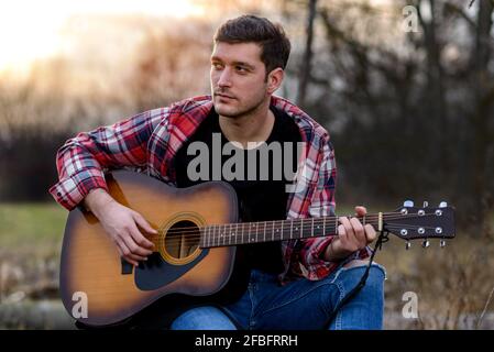 Ein hübscher junger Mann spielt bei Sonnenuntergang im Naturpark akustische Gitarre. Stockfoto