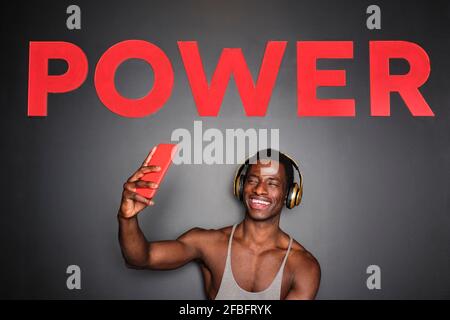 Fröhlicher junger Mann mit Kopfhörern, der Selfie unter dem Powertext macht An der Wand im Fitnessstudio Stockfoto