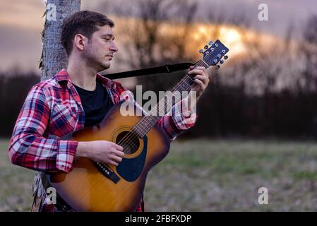 Ein hübscher junger Mann, der sich gegen einen Baum lehnt, spielt bei Sonnenuntergang in einem Naturpark eine akustische Gitarre. Stockfoto