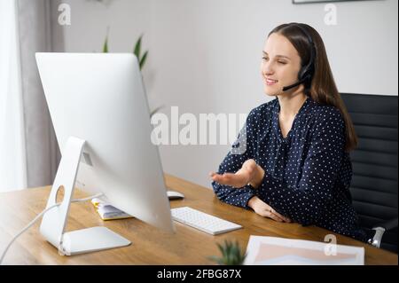 Eine positive Frau ist eine Kundendienstmitarbeiterin, die am Schreibtisch sitzt und den Kunden online befragt, eine Mitarbeiterin, die ein Headset trägt, spricht mit Kollegen aus der Ferne, beteiligt sich an einem Videomeeting Stockfoto