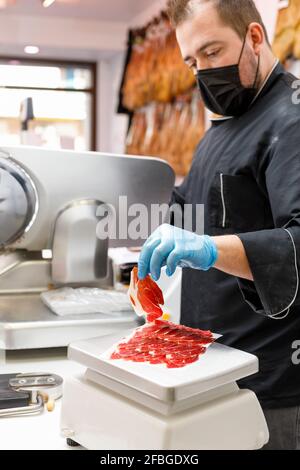 Kaukasischer Metzger mit iberischem Schinken auf Waage im Geschäft während Pandemie Stockfoto