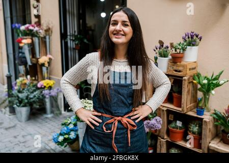 Lächelnde junge Floristin, die mit den Händen auf den Hüften steht Blumenladen Stockfoto