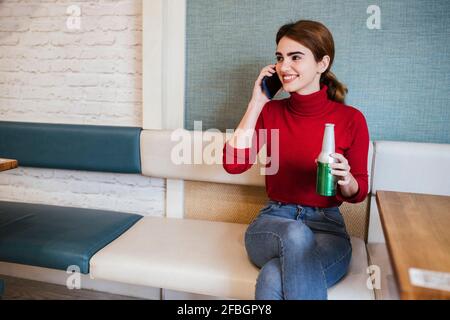 Lächelnde Frau mit Bierflasche, die beim Gespräch wegschaut Handy in der Bar Stockfoto