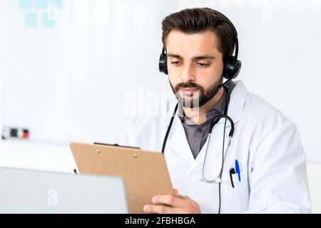 Hübscher männlicher Arzt, der Kopfhörer trägt, während er Bericht auf der Zwischenablage liest Im Krankenhaus Stockfoto