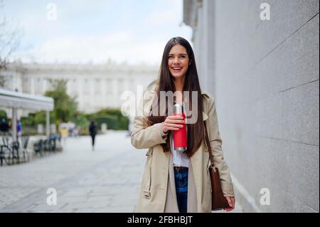 Aufgeregte Frau, die einen isolierten Getränkebehälter in der Nähe der Wand hält In der Stadt Stockfoto