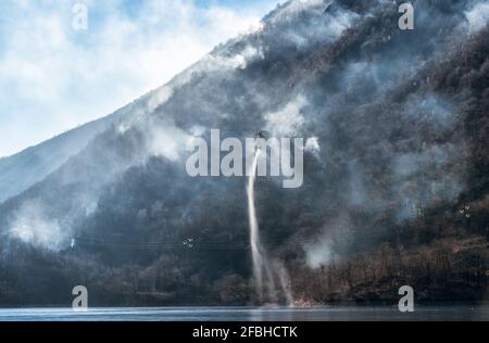 Feuer des Mondonico-Gebirges über dem Ghirla-See mit einem Hubschrauber, der Wasser zum Löschen einlädt, Valganna, Lombardei, Italien. Stockfoto