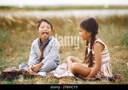 Mädchen, das Bruder mit Down-Syndrom betrachtet, der aus der Zunge ragt, während er auf dem Gras sitzt Stockfoto