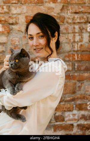 Schöne Frau, die die Katze Chartreux vor einer Ziegelmauer hält Stockfoto