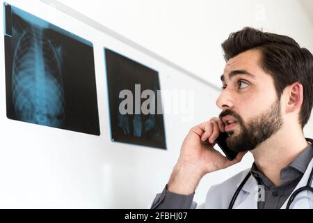 Männlicher Arzt, der Röntgenaufnahmen ansieht, während er auf dem Mobiltelefon spricht Stockfoto