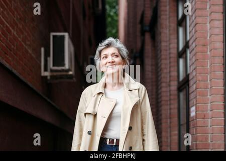 Lächelnde reife Frau, die beim Gebäude steht und wegschaut Stockfoto