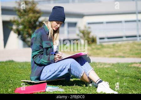 Junge Frau, die an sonnigen Tagen auf dem Skateboard auf dem College-Campus sitzt, studiert Stockfoto