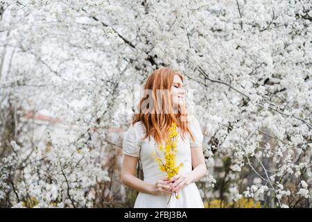 Rothaarige junge Frau, die im Stehen einen Strauß gelber Blume hält Vor dem Mandelbaum Stockfoto