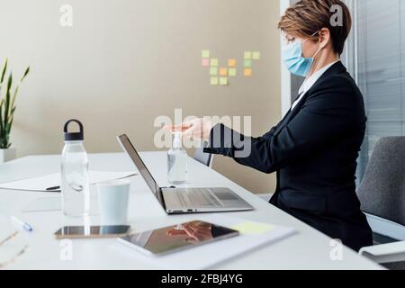 Weibliche Unternehmerin mit schützender Gesichtsmaske mit Handdesinfektionsmittel, während Am Schreibtisch sitzend Stockfoto