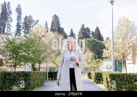 Lächelnde, reife Frau, die mit einem wiederverwendbaren Becher davonschaut Auf Fußweg Stockfoto
