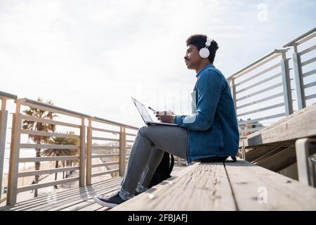 Ich betrachtete einen Mann, der Smartphone über den Laptop hält, während er auf dem Laptop sitzt Schritte an sonnigen Tagen Stockfoto