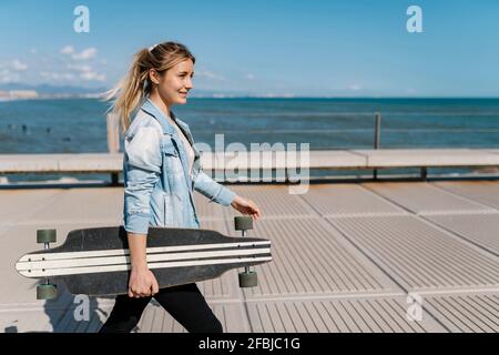 Lächelnde Frau mit Longboard, die an sonnigen Tagen auf dem Pier läuft Stockfoto