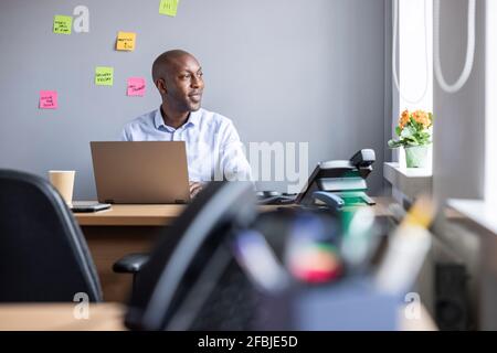 Ein nachdenklicher Geschäftsmann schaut weg, während er mit einem Laptop im Büro sitzt Stockfoto