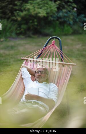 Reifer Mann mit Buch, der sich in der Hängematte im Hinterhof entspannt Stockfoto