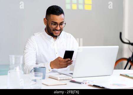 Junger Geschäftsmann, der Smartphone am Laptop am Schreibtisch benutzt Büro Stockfoto