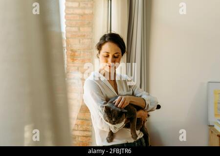 Lächelnde Frau streichelte die Katze vor dem Fenster zu Hause Stockfoto