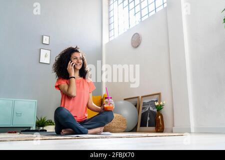 Lächelnde Frau, die mit dem Handy spricht, während sie das Saftglas in der Hand hält Zu Hause Stockfoto